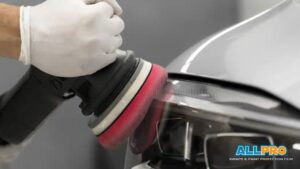 A detailer wearing gloves using a red foam buffer to polish the headlight area of a silver car with the “AllPro Wraps & Paint Protection Film” logo in the corner