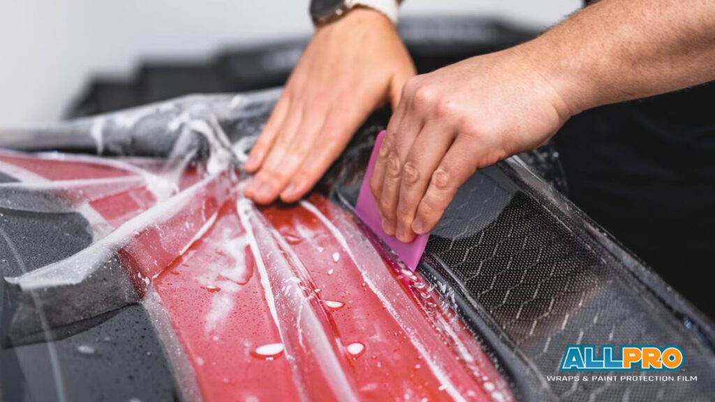 A close-up of hands using a squeegee to smooth protective film on a red car hood with carbon fiber accents (Paint Protection Film PPF)