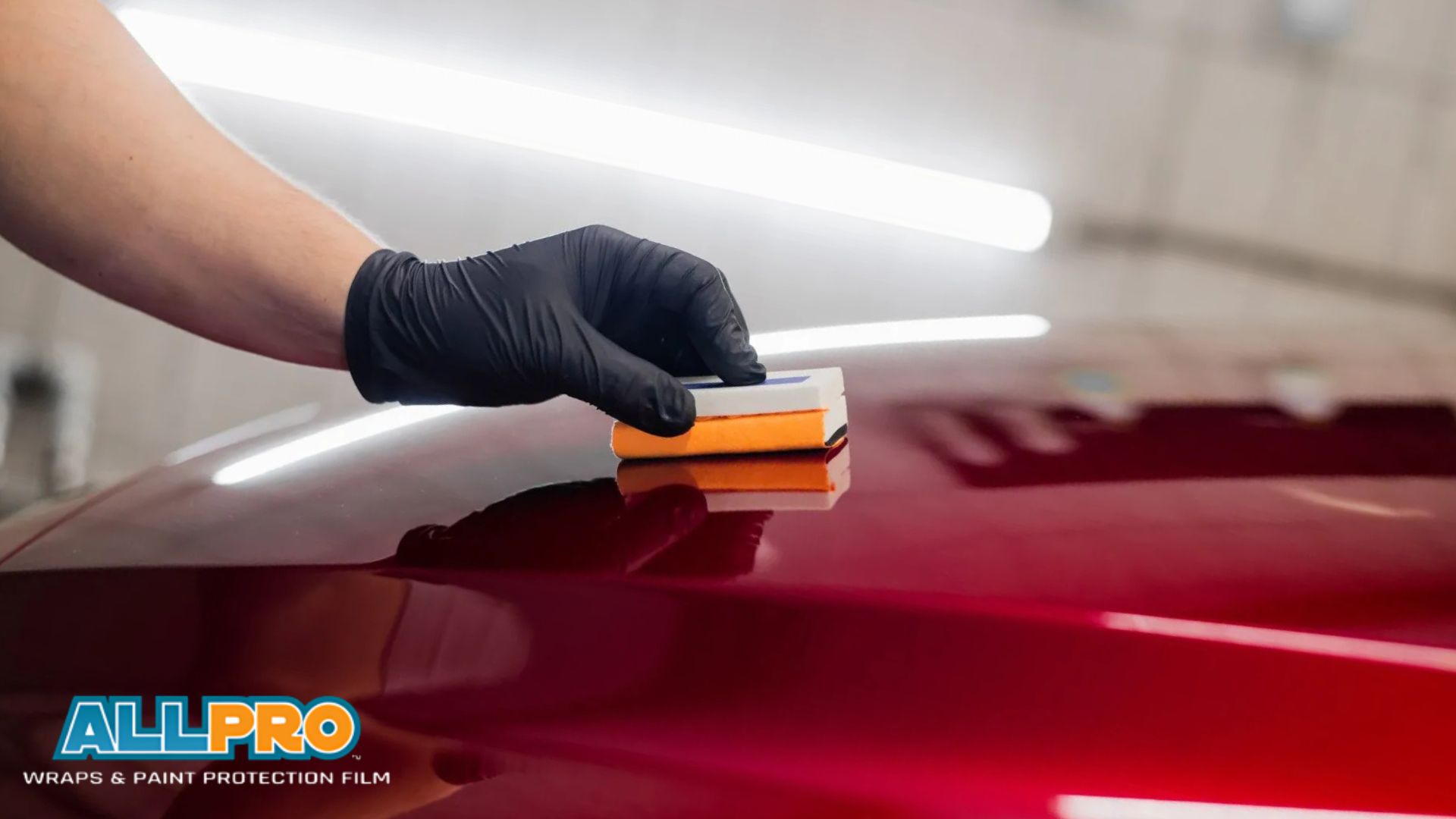 A close-up of a gloved hand applying paint protection film on a glossy red car surface using a small applicator. The logo "All Pro Wraps & Paint Protection Film" is visible in the corner