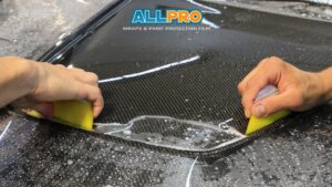 Two hands using yellow squeegees to apply protective film on a carbon fiber car hood covered in water droplets