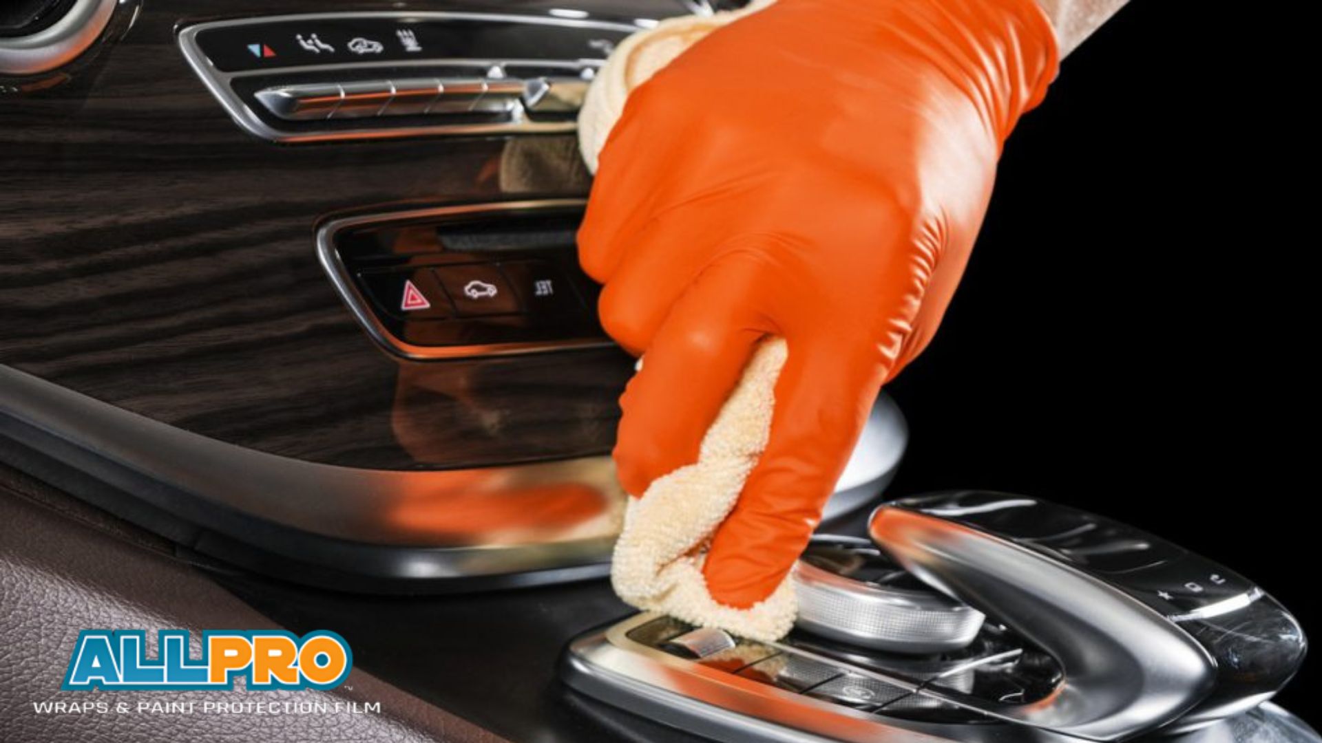 A close-up of a gloved hand in orange latex cleaning the glossy center console of a luxury car with a soft beige cloth. The surface includes buttons and sleek wood paneling