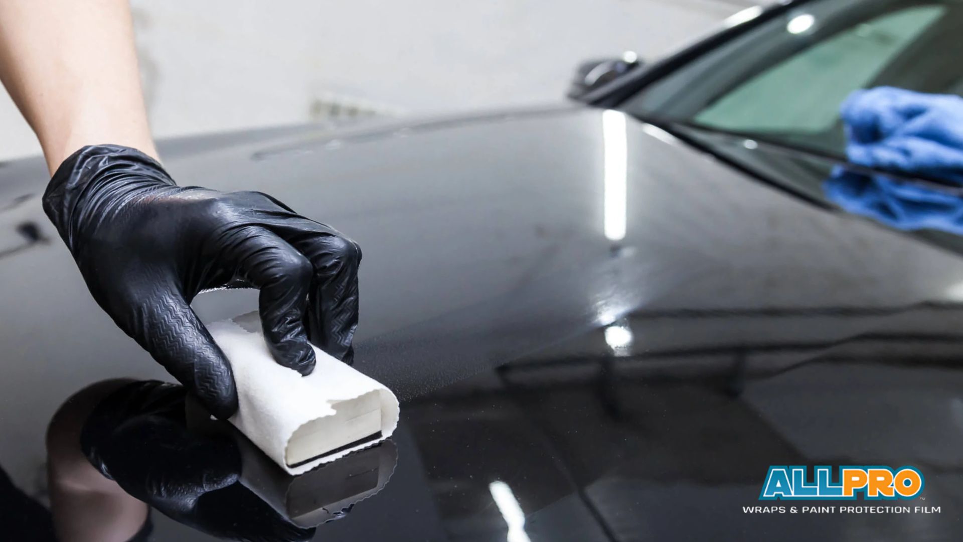 A professional detailer wearing black gloves applies a ceramic coating or protective film to a black car's hood using an applicator pad.