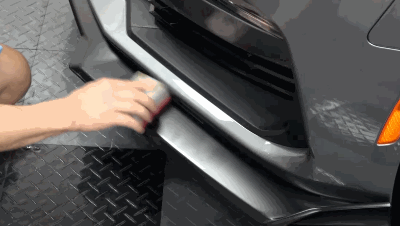 A person applies detailing product to a car’s silver front bumper using an applicator pad, enhancing the finish around the grille and lower lip.