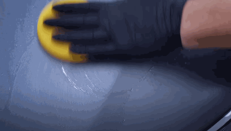 A gloved hand uses a yellow foam pad to apply polish in circular motions to a dark-colored car hood, bringing out the shine.