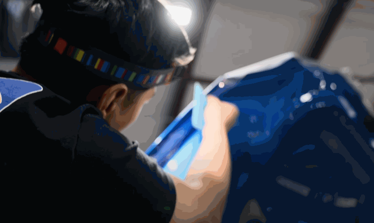 A technician in a headlamp uses a blue squeegee to apply paint protection film to the hood of a blue vehicle, focusing on edge work.