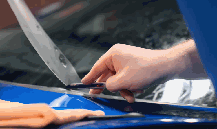 A close-up of a hand trimming the excess paint protection film with a precision blade near a blue car's windshield.