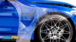 A close-up of a blue BMW with paint protection film being installed, showing the film partially laid over the fender and wheel arch.
