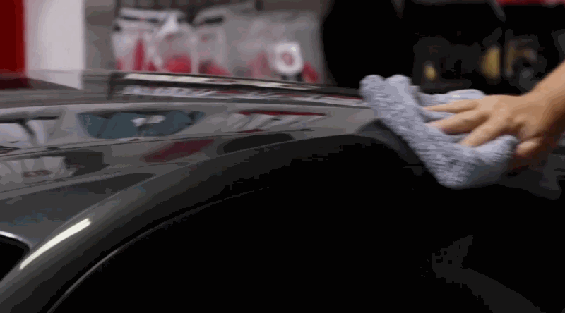 A hand wipes down a black car roof with a plush microfiber towel, removing dust and residue to prepare for paint protection film installation.