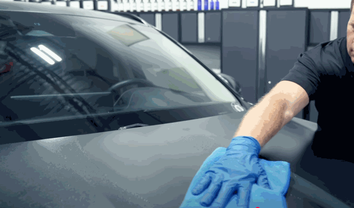 A man in a black shirt, wearing blue gloves, carefully buffs the hood of a black vehicle in a well-lit garage using a microfiber towel.