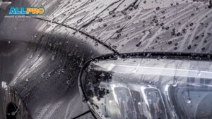 A close-up of a car's headlight and hood covered in water beads after rain, highlighting the glossy, water-repellent surface protected by paint protection film.