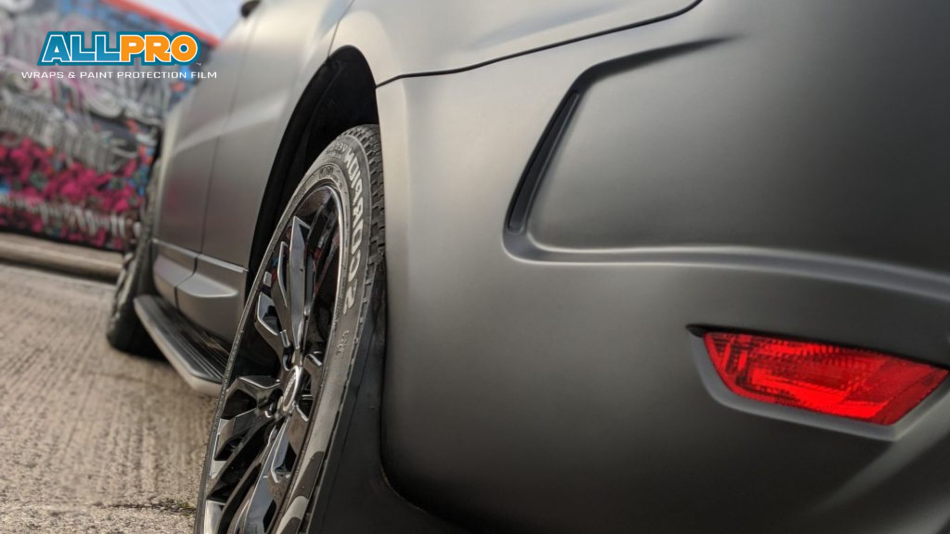 Close-up of a matte grey SUV’s rear wheel and bumper with glossy black rims, featuring the AllPro Wraps & Paint Protection Film logo in the top left corner and a graffiti-covered vehicle blurred in the background.