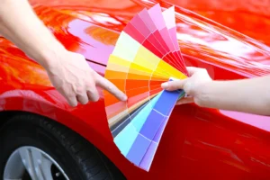 A person holds a wide fan of vinyl wrap color samples next to a shiny red car, pointing to an orange-red shade for comparison.