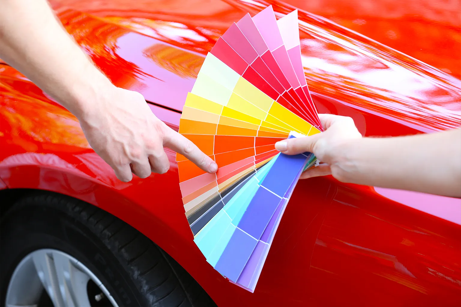 A person holds a wide fan of vinyl wrap color samples next to a shiny red car, pointing to an orange-red shade for comparison.
