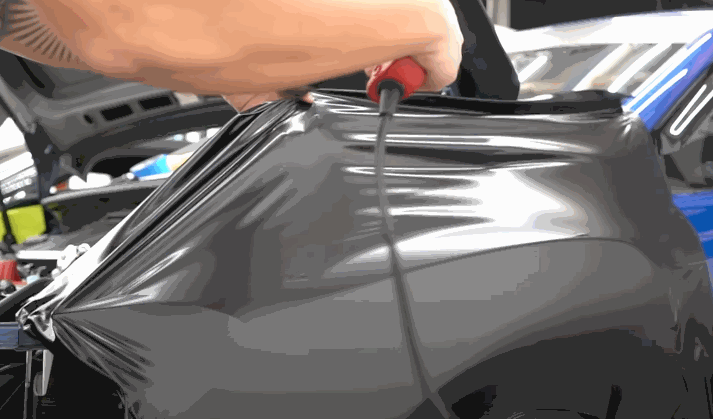 A person uses a heat gun to stretch and smooth glossy black vinyl wrap over a car's body panel.