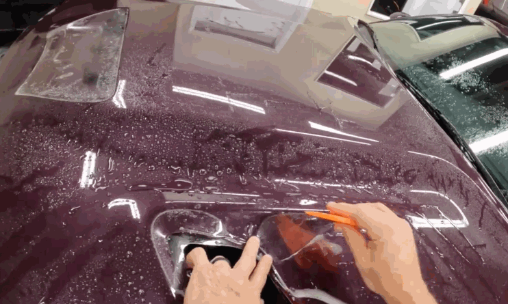 Applying paint protection film: A person uses a squeegee to smooth a transparent protective film over the hood of a burgundy car, ensuring proper adhesion for paint protection.