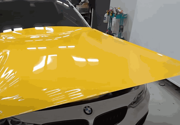 A technician applying a large sheet of yellow vinyl wrap to the hood of a BMW, smoothing it out for an even finish.