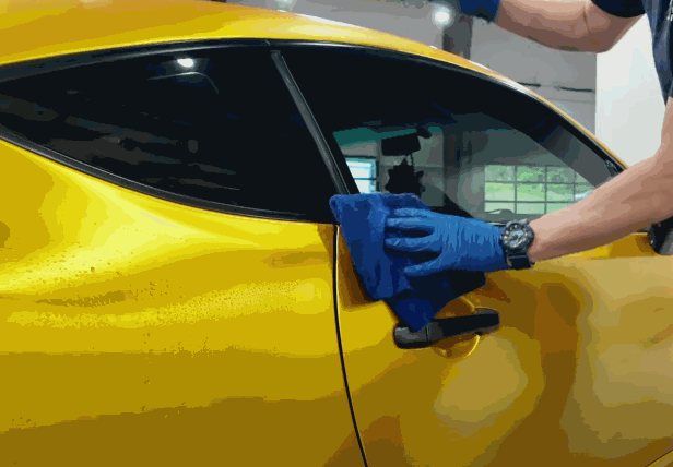A person wiping down a yellow car's surface with a microfiber cloth to prepare it for the vinyl wrap application.