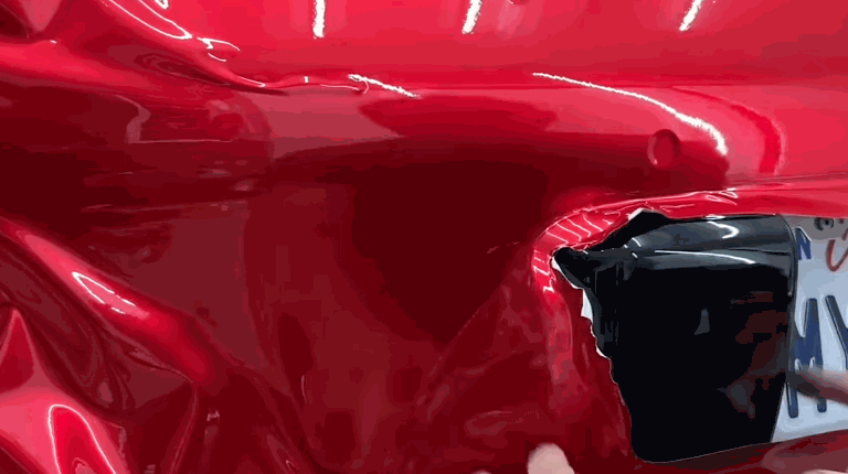 Red vinyl wrap being carefully molded around the curved edges of a car’s rear panel near the license plate.