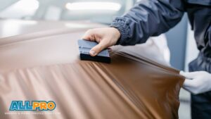 Close-up of a technician using a felt-edge squeegee to apply a satin brown vinyl wrap smoothly across a car surface, with the AllPro Wraps & Paint Protection Film logo in the corner.