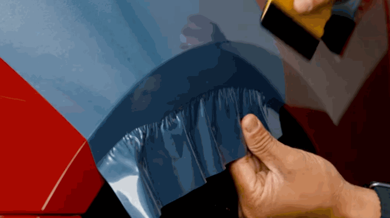 Close-up of a person applying a blue vinyl wrap to a red vehicle using a squeegee to smooth out air bubbles.