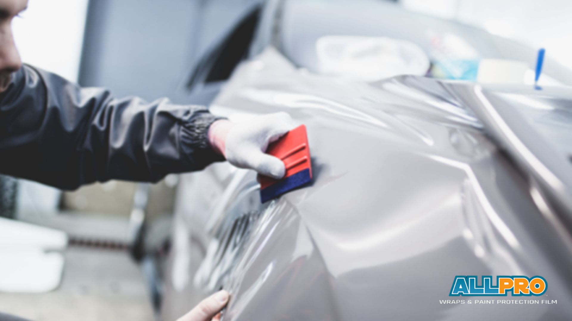 Installer smoothing grey vinyl wrap onto a car with a red and blue squeegee tool, wearing gloves for precision. The logo “ALLPRO Wraps & Paint Protection Film” is visible in the corner.