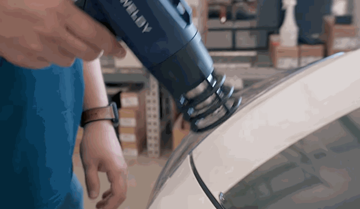 Vinyl wrap removal step showing a heat gun being used on the rear panel of a white car to loosen the film for safe detachment.