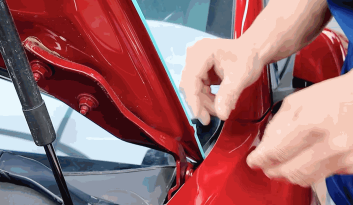 Close-up of vinyl wrap removal from a red car near the hood hinge, with hands carefully pulling back the blue wrap from the tight edge.