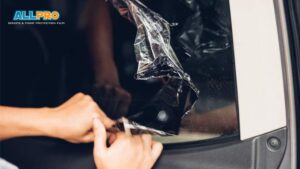Close-up of vinyl wrap removal from a car window, showing hands carefully peeling off a dark, glossy tint film. The wrap curls as it’s pulled back, revealing the clear glass underneath.
