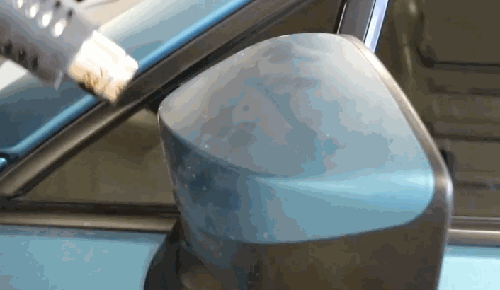 A heat gun is applied to a blue car's side mirror during vinyl wrap removal, softening the adhesive to make peeling the wrap easier and cleaner.