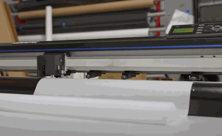 A precision vinyl cutter machine slicing through a sheet of protective film in a workshop setting, with rolled materials visible in the background.