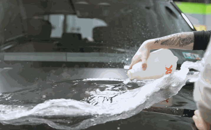 A person with tattooed arms sprays a liquid solution over a transparent film on a vehicle’s hood, smoothing it into place for paint protection.