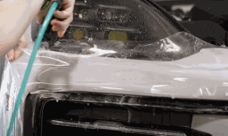 Hands stretch and position clear paint protection film across the front bumper and headlight of a vehicle, using water and tools for alignment.