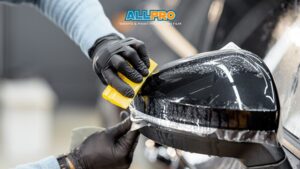 A close-up of a technician in black gloves using a yellow squeegee to apply clear paint protection film to a car’s side mirror. The ALLPRO Wraps & Paint Protection Film logo is visible above the action.