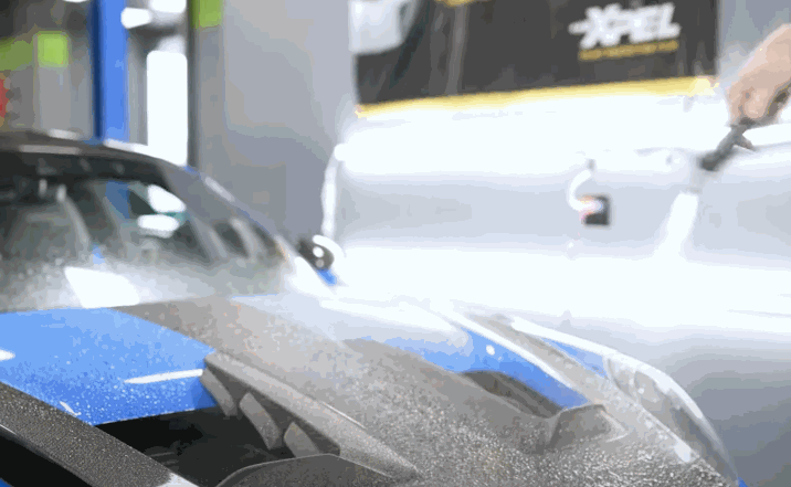 A close-up shot of a technician spraying a solution onto the surface of a blue sports car, prepping it for paint protection film installation.