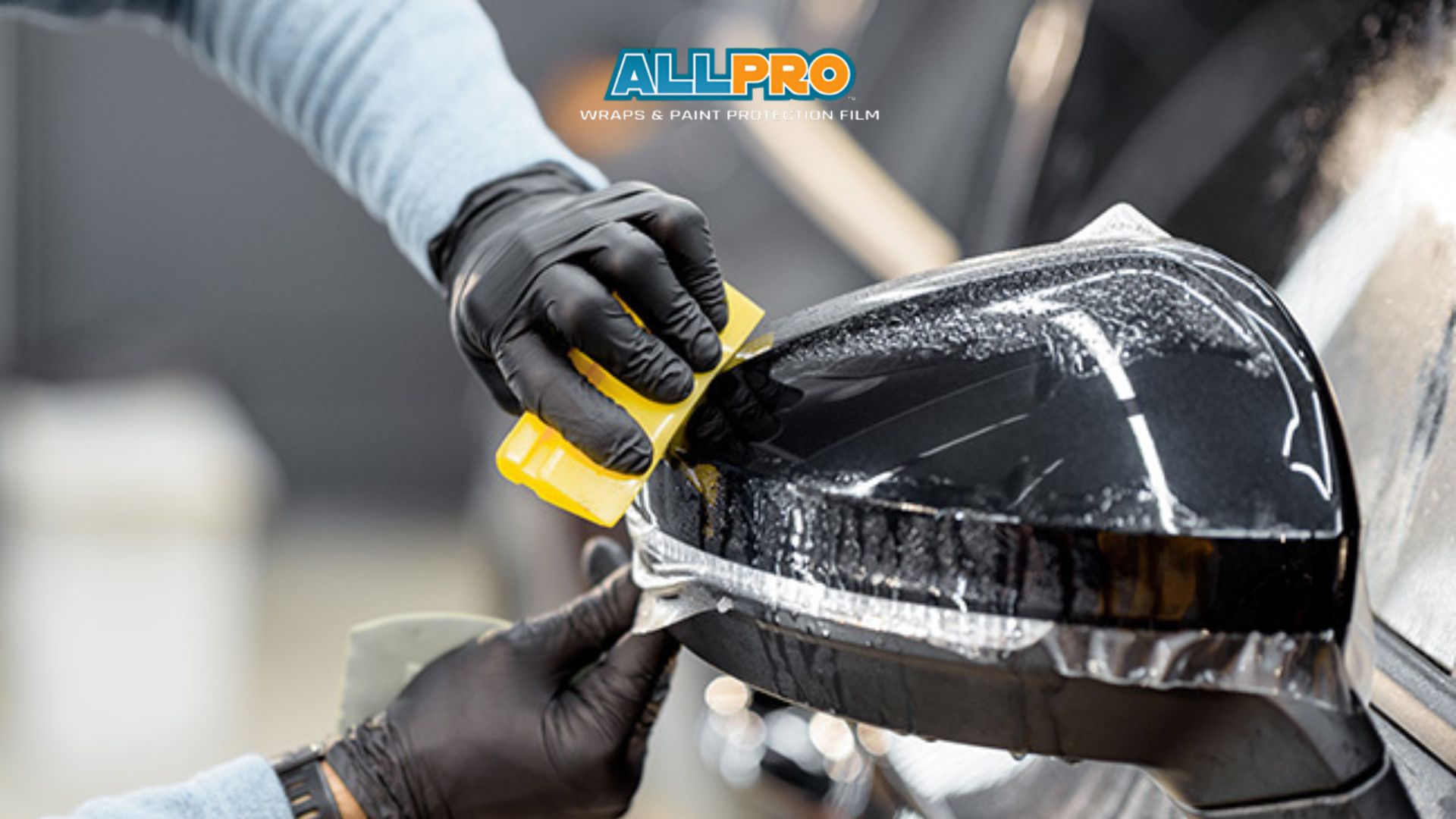 A close-up of a technician in black gloves using a yellow squeegee to apply clear paint protection film to a car’s side mirror. The ALLPRO Wraps & Paint Protection Film logo is visible above the action.