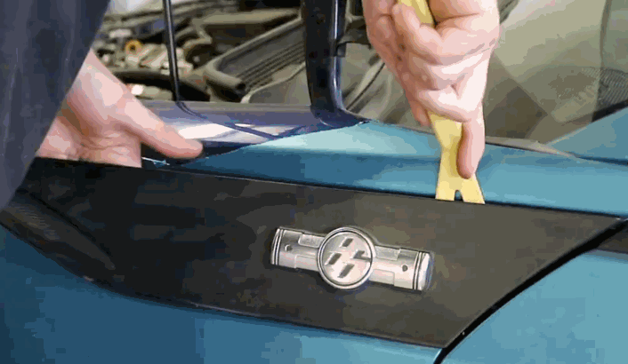 Vinyl wrap removal in progress as a person uses a yellow pry tool to lift trim near the side mirror of a blue car, exposing edges of the wrap beneath the black panel.