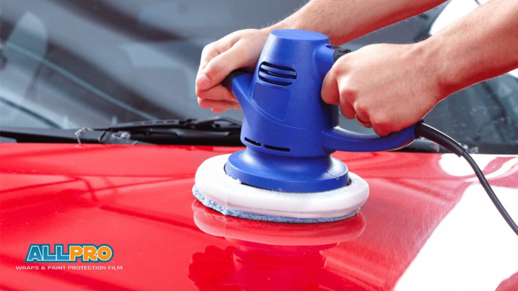 Close-up of a person using a blue dual-action polisher on a red car hood to remove ceramic coating.