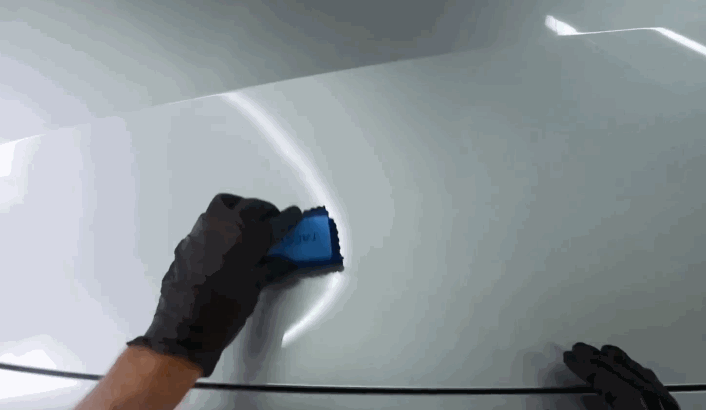 A gloved hand wiping a smooth car hood with a blue applicator pad during ceramic coating removal.