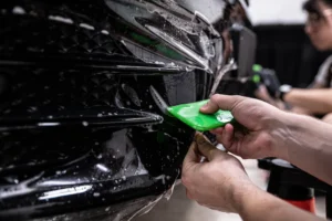 Installer uses a green squeegee to apply paint protection film to the front bumper of a black vehicle at AllPro Wraps.