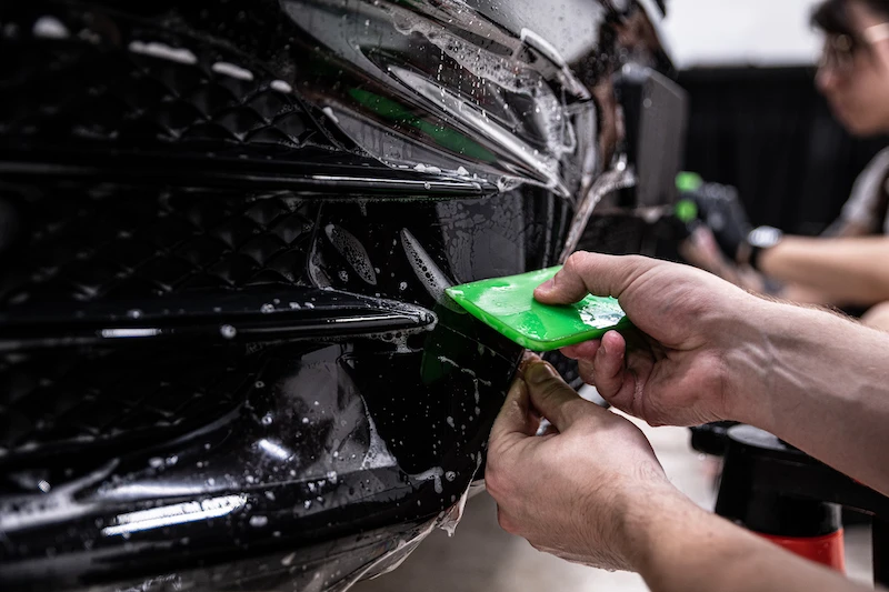 Installer uses a green squeegee to apply paint protection film to the front bumper of a black vehicle at AllPro Wraps.