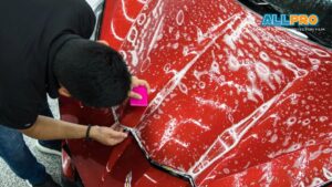 An installer using a pink squeegee to press paint protection film onto the hood of a red sports car, with soapy water covering the surface.