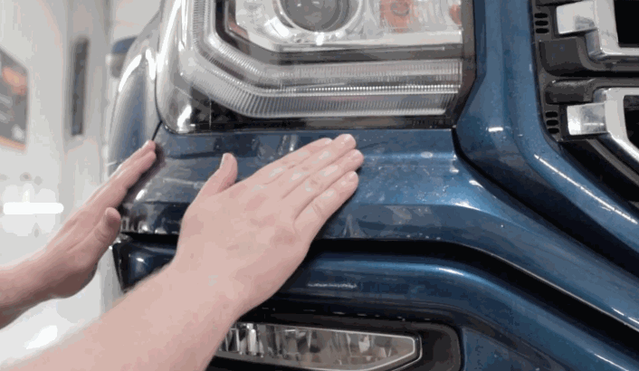 Hands smoothing paint protection film onto the front bumper and headlight area of a blue truck.