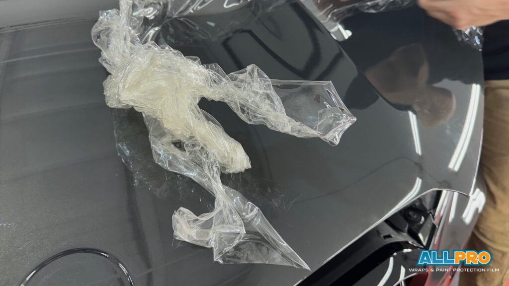 Crinkled paint protection film (PPF) partially removed from a glossy dark car hood, with a technician's hand visible in the background.