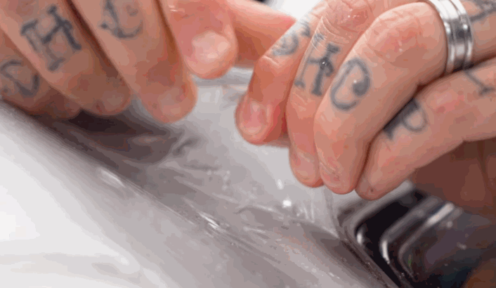 Tattooed hands carefully stretch and apply clear PPF over a white car surface, focusing on smooth adhesion around a vent.