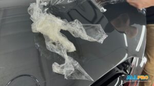 Crinkled paint protection film (PPF) partially removed from a glossy dark car hood, with a technician's hand visible in the background.