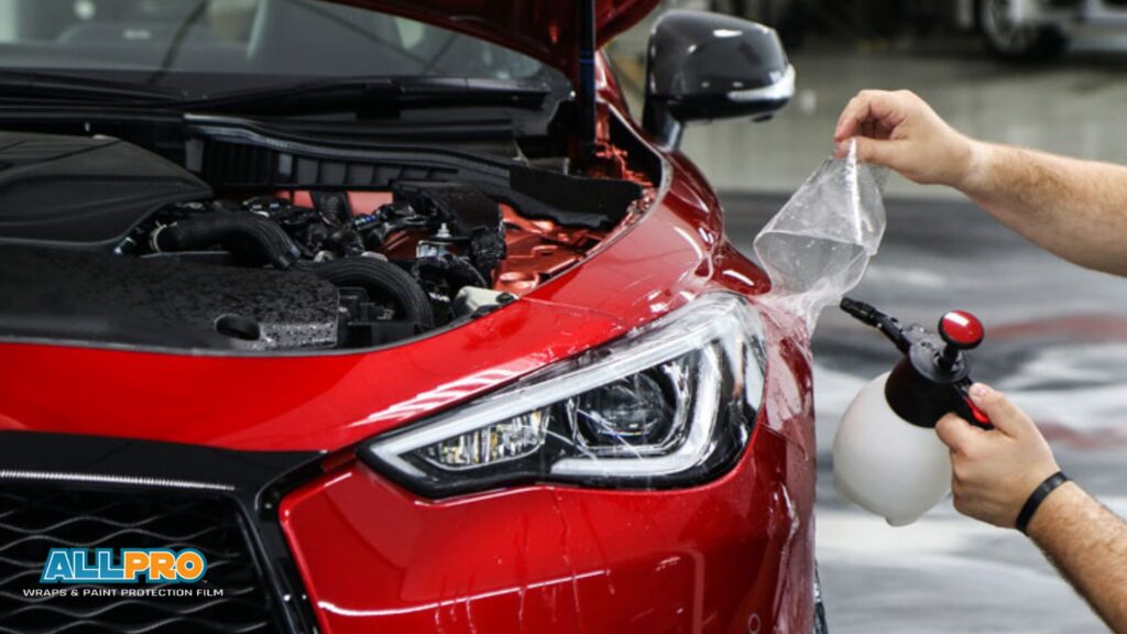 Close-up of a red car with its hood open as a technician applies paint protection film to the front fender using a spray bottle.
