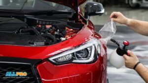 Close-up of a red car with its hood open as a technician applies paint protection film to the front fender using a spray bottle.