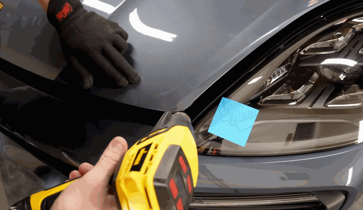 Technician using a heat gun to apply paint protection film on the front edge of a gray car’s hood while wearing gloves for precision.