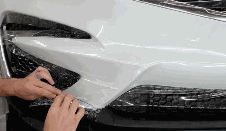 Hands carefully stretch and smooth Paint Protection Film across the front bumper of a white vehicle. The clear film is aligned around the grille and body contours to provide full surface protection.