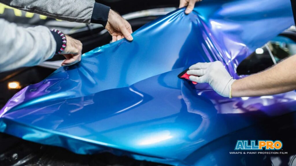 Technicians applying blue and purple vinyl wrap to a car hood using a squeegee to smooth the material and remove air bubbles during installation.